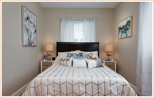 Interior of bedroom. Bed with white and gold comforter and pillows in front of window.